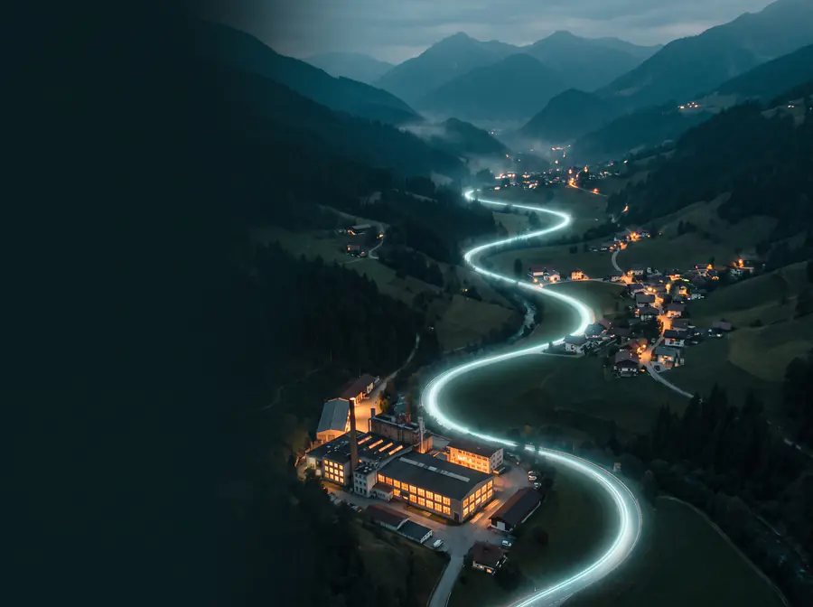 Ein Alpental zur blauen Stunde, eine Serpentinenstrasse, an der ein Strom aus Licht entlangfliesst, vorbei an einer Fabrik und Dörfern.
